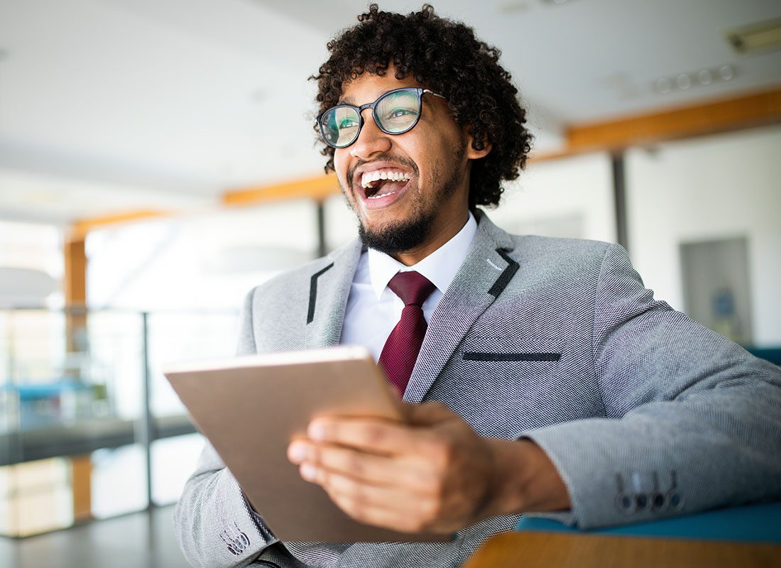 Contact - Closeup Portrait of a Cheerful Young Businessman Wearing a Suit and Tie Sitting on a Chair in the Office While Holding a Tablet in his Hands