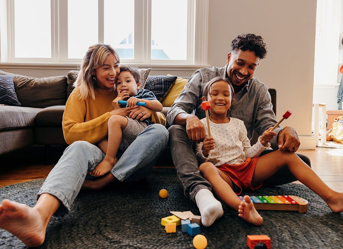Personal Insurance - Portrait of Two Cheerful Young Parents Having Fun Playing with Their Two Kids in the Living Room While Sitting on the Rug